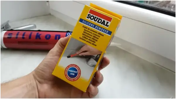 Ręka trzymająca opakowanie środka do usuwania silikonu Soudal. W tle widać tubkę z silikonem.