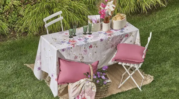 Un tavolo da giardino apparecchiato con una tovaglia floreale, bicchieri verdi, cesti e cuscini rosa. Un'idea per come rivestire un tavolo con stoffa per un picnic.