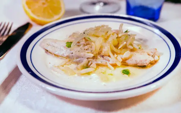 Delicato branzino in padella con cipolle e prezzemolo, servito su un piatto bianco con bordo blu. Accanto, uno spicchio di limone.