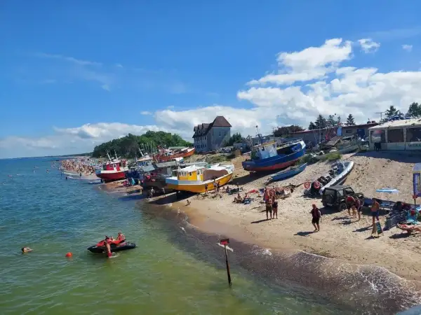 Słoneczny dzień nad polskim morzem. Kolorowe łodzie na plaży, tłumy ludzi, a na wodzie skuter wodny.