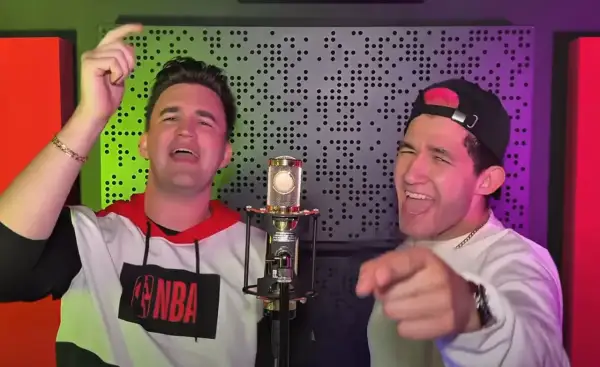 Dwóch mężczyzn śpiewa do mikrofonu. Jeden ma na sobie bluzę z logo NBA, drugi czapkę z daszkiem. W tle widać panel akustyczny.