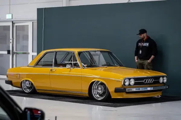 Gul Audi 100 C1, en klassiker med kromade fälgar, står parkerad bredvid en man i keps.