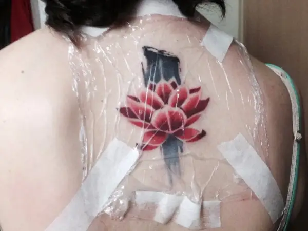 Flor de loto roja y azul recién tatuada en la espalda, cubierta con plástico y cinta. Preguntándose cuanto tarda en cicatrizar un tatuaje.