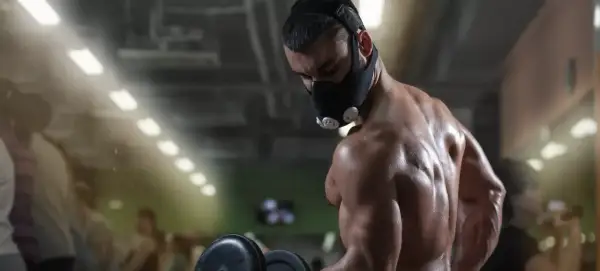 A muscular man performs dumbbell curls in a gym, wearing an oxygen mask for altitude training.
