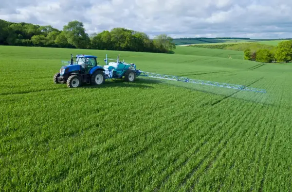 Niebieski traktor z opryskiwaczem na zielonym polu, stosuje środek Chwastox Turbo 340 SL. Dawkowanie jest kluczowe dla skuteczności.