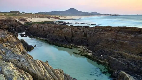 Aguas cristalinas en la **playa das Furnas**, entre rocas volcánicas y arena dorada, con el mar al fondo y una montaña.