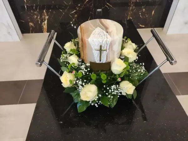 Białe róże i gipsówka otaczają urnę z krzyżem. Kwiaty na pogrzebie z urną symbolizują pamięć i szacunek.