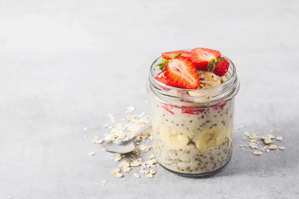 En burk med overnight oats recept toppad med jordgubbar och banan. Havregryn och en sked ligger bredvid.