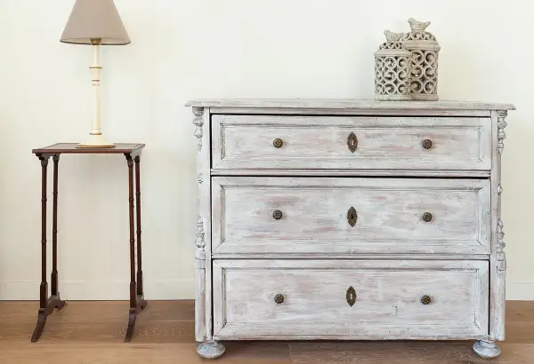 Un meuble cérusé patiné, avec ses tiroirs et ses ornements délicats, se dresse à côté d'une petite table d'appoint surmontée d'une lampe.