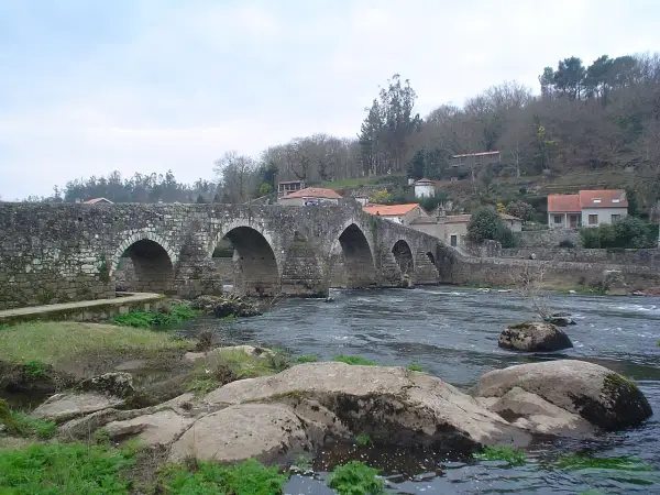 El ponte Maceira, un puente de piedra con arcos, cruza un río caudaloso. Casas con tejados rojos se asoman en la ladera.