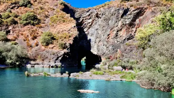 Un río de aguas turquesas fluye hacia un monte furado, una cueva natural en la roca, con vegetación y personas disfrutando del paisaje.