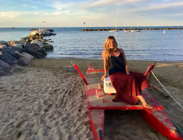 Donna sorridente su una barca rossa sulla spiaggia di Cattolica, Emilia Romagna, con il mare e i faraglioni sullo sfondo.