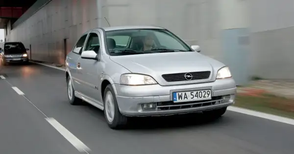 Srebrna astra 2 jedzie drogą, światła włączone, kierowca za kierownicą.