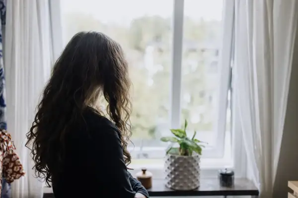 Flicka med långt hår tittar ut genom fönstret. En planta står på fönsterbrädan. Bilden kan symbolisera emotionell utveckling 3 år.
