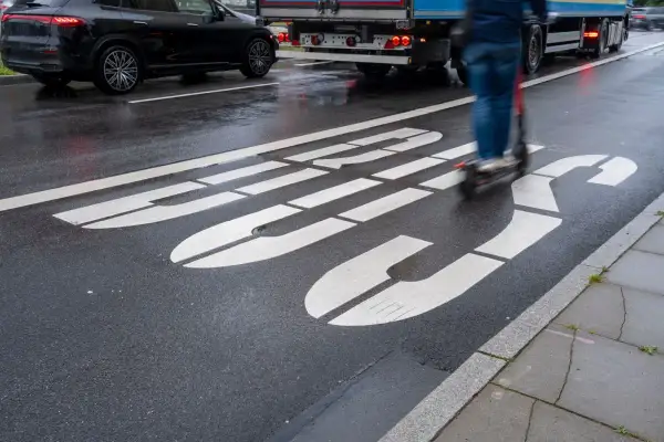 Jazda hulajnogą po buspasie grozi mandatem. Na mokrej drodze widać białe linie wyznaczające pas oraz poruszającego się po nim użytkownika hulajnogi.