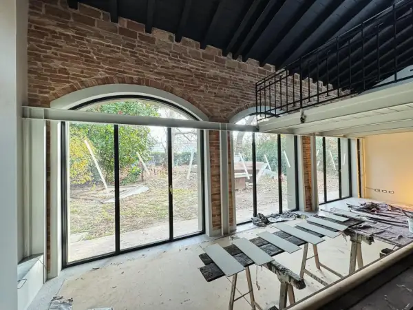 Interno in fase di ristrutturazione con ampie vetrate sul giardino. In primo piano, assi di legno appoggiate su cavalletti, segno di lavori di miglioramento della casa in corso.