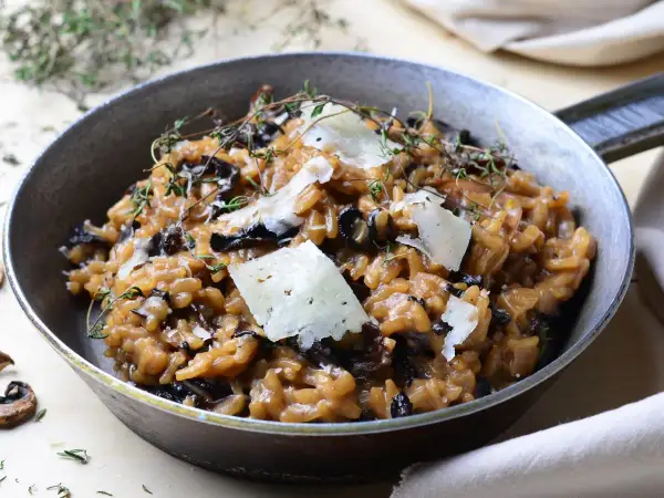 Aksamitne risotto z grzybami, udekorowane płatkami parmezanu i gałązkami tymianku, podane w metalowej patelni.
