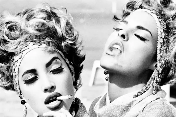 Iconica **Helmut Newton moda**: due donne con acconciature voluminose e make-up audace fumano sigarette, un'immagine di stile e provocazione.