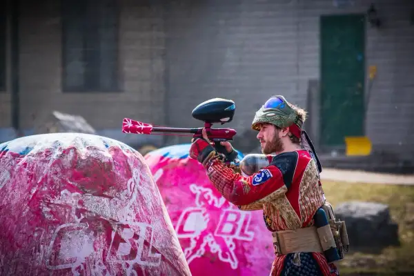 Gracz w stroju bojowym celuje z markerem, gotowy do akcji w laserowym paintballu. Za nim różowe dmuchane przeszkody.