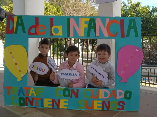 Niños sonríen tras un marco decorado para el Día de la Infancia, sosteniendo carteles con palabras como "Juego", "Familia" y "Protección".
