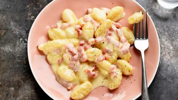 Ñoquis a la carbonara cremosos y deliciosos, cubiertos con trozos de panceta y pimienta negra, servidos en un plato rosa.