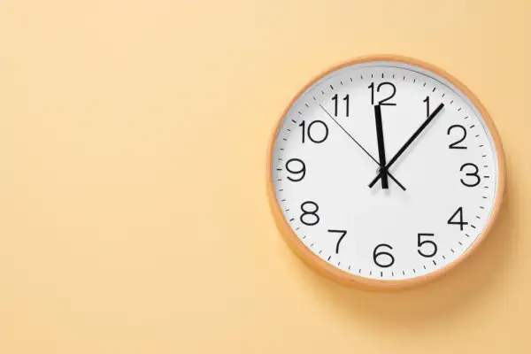 Reloj de pared blanco con marco de madera. Las manecillas marcan las 12:05. Ideal para aprender las horas en inglés, ejemplos.