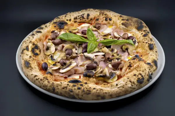 Pizza Capricciosa con champiñones, jamón y aceitunas negras, coronada con albahaca fresca. ¡Un verdadero capricho!