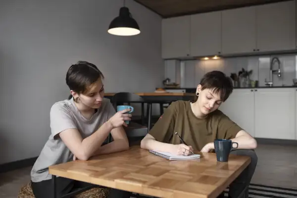 Dwie osoby przy stole, jedna pisze, druga pije kawę. Rozmowa o tym, jak załatwić zaświadczenie o dysleksji, może być wsparciem w nauce.