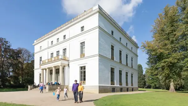 Familie spaziert durch den Jenisch Park vor einem weißen Herrenhaus mit Säulen und goldenen Verzierungen.
