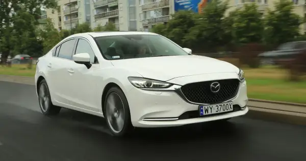 Biała Mazda 6 2.0 benzyna jedzie po mokrej drodze, w tle budynki i drzewa.