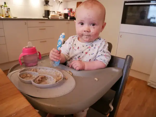 Sött litet barn med mat i ansiktet, redo att introducera mat bebis. En tallrik med mat och en mugg finns bredvid.