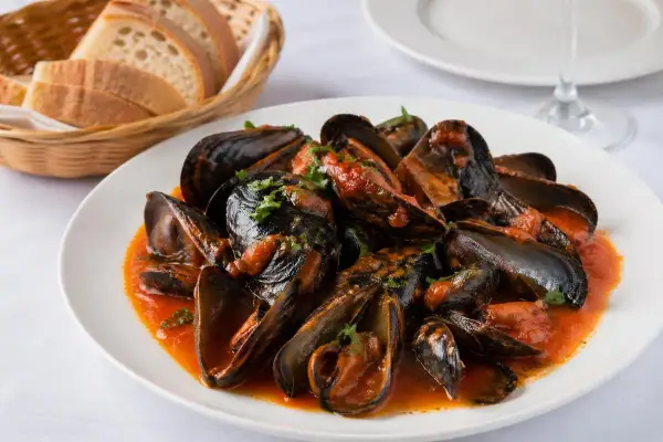 Mejillones a la gallega servidos en un plato blanco con salsa de tomate y perejil picado. Al fondo, una cesta de pan y una copa de vino.