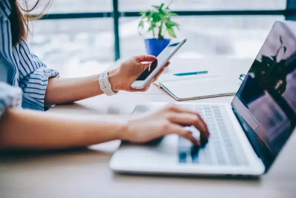 Kobieta trzyma telefon, planując, jak udostępnić internet z telefonu na komputer. Pracuje przy laptopie.