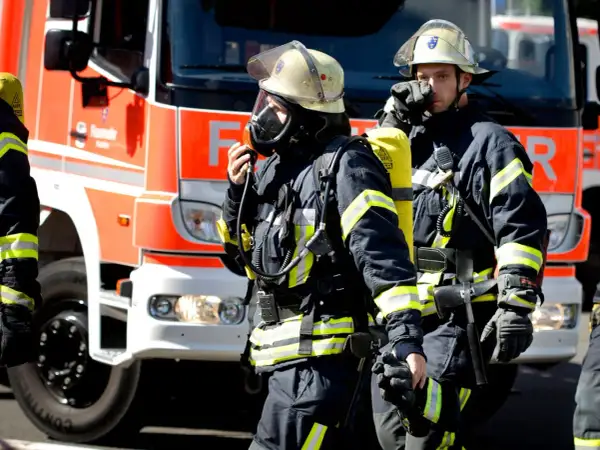 Zwei Feuerwehrmänner in voller Montur vor einem roten Feuerwehrauto. Sie sind bereit für den Einsatz. Wie viel verdient man als Feuerwehrmann?