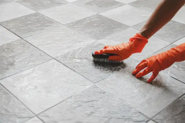 Mani con guanti arancioni spazzolano le fughe del pavimento con schiuma, mostrando come pulire le fughe del pavimento per un risultato brillante.
