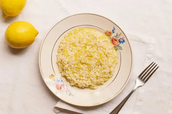 Un cremoso risotto al limon, con ralladura de limón espolvoreada, servido en un plato floral. Dos limones frescos reposan cerca.