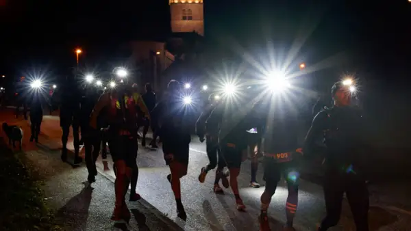 Courseurs de trail de nuit, lampes frontales allumées, traversant un village sombre. Un chien les accompagne.