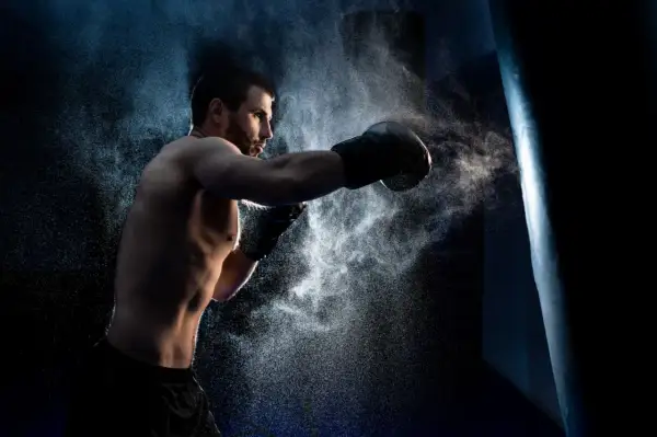 A determined boxer, embodying the characteristics of a fighter, throws a punch at a heavy bag, kicking up a cloud of dust.