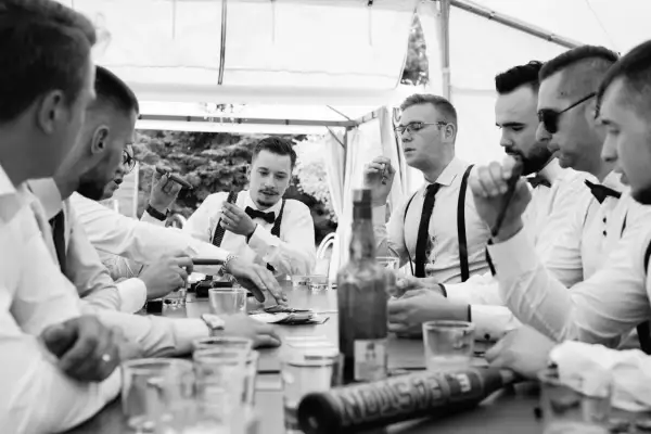 Panowie w eleganckich koszulach i muszkach świętują kawalerski przy stole z cygarami i butelką whisky.
