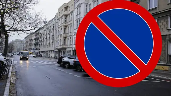 Znak B35 zakazu zatrzymywania się na mokrej ulicy z samochodami i kamienicami w tle.
