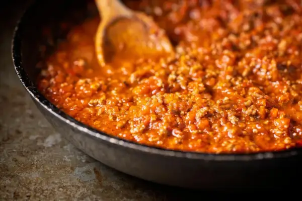 Herzhafte bolognese sauce köchelt in einer Pfanne, bereit für Pasta. Ein Holzlöffel rührt die fleischige Tomatensauce.