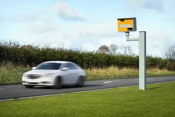 Biały samochód pędzi drogą, a obok stoi fotoradar. Chyba niedługo przyjdzie mandat z fotoradaru.