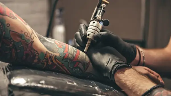 Manos enguantadas trabajan en un tatuaje colorido en un brazo. El artista usa una máquina de tatuar, creando arte permanente.