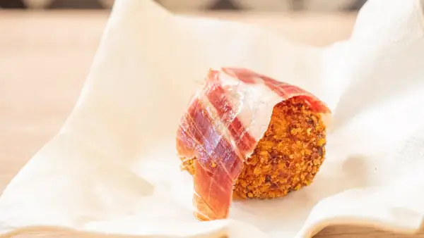 Croquetas de jamón crujientes, envueltas en una loncha de jamón serrano, servidas sobre papel blanco.