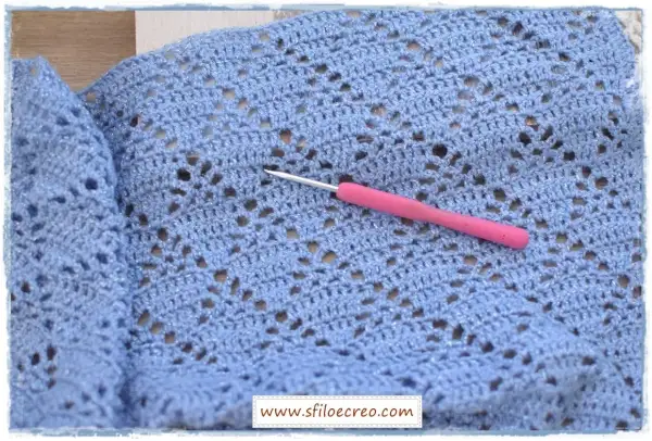 Un uncinetto rosa riposa su un lavoro a maglia azzurro con fili scintillanti, mostrando un delicato punto incrociato uncinetto.
