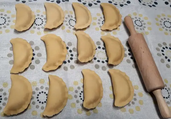 Gotowe do gotowania pierogi z jabłkami, ułożone rzędami na wzorzystym obrusie, obok drewnianego wałka.
