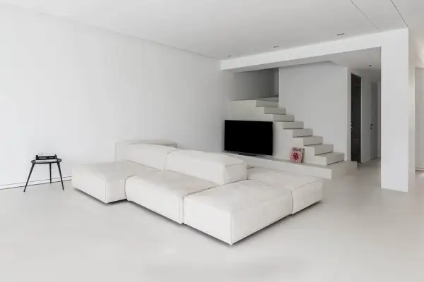 Soggiorno minimalista con divano modulare bianco e pavimento in resina epossidica opaca. Scale moderne conducono a un'area con TV.