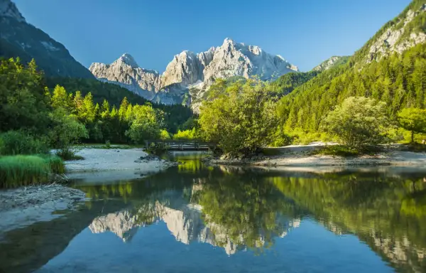 En vacker vy för att vandra i Slovenien. En spegelblank sjö reflekterar berg och skog. En bro leder över vattnet.