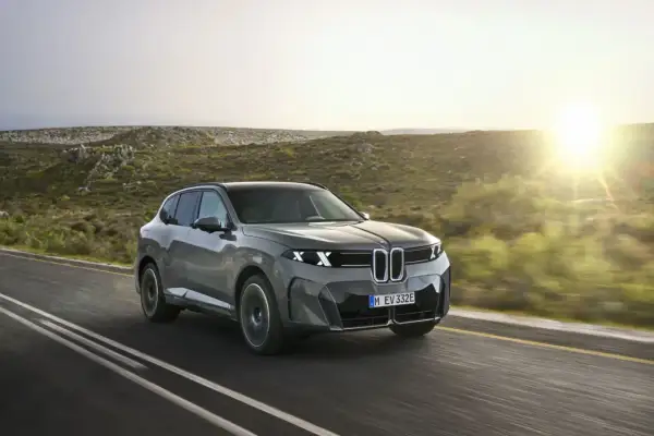 El BMW iX5, un SUV eléctrico gris, avanza por una carretera rural al atardecer, con colinas verdes y el sol brillando.