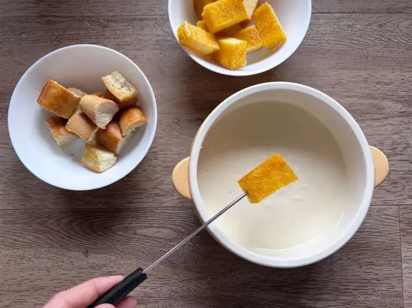 Una fetta di pane croccante viene intinta in una cremosa fonduta di parmigiano, accanto a ciotole di crostini.